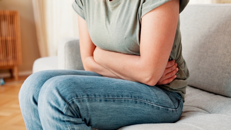 Žena sedi savijena na kauču i drži stomak zbog bola