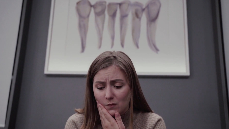 Žena sedi zabrinuto u stomatološkoj ordinaciji i drži obraz zbog jakog bola