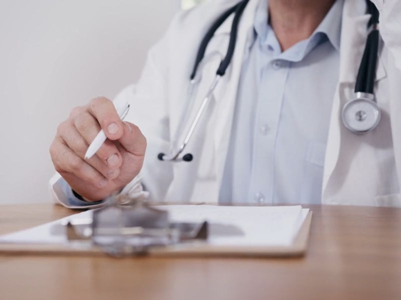 prikaz lekara koji sedi za stolomi drži hemijsku u ruci, ispred njega kartonska lista